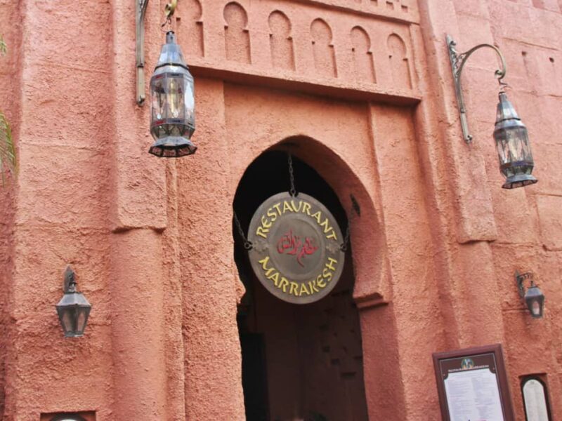 Entrée du restaurant Marrakesh à Disney World Orlando, avec une enseigne ronde suspendue et deux lanternes, sur fond de murs texturés rose-orange et d'arcades ornées dans le pavillon du Maroc.