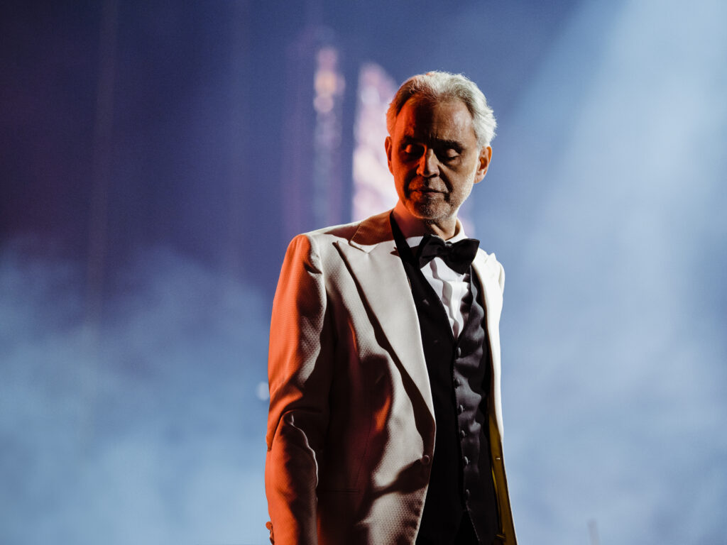 Un homme vêtu d'une veste de smoking blanche et d'un nœud papillon noir se tient sur scène sous un éclairage dramatique lors d'un concert au Vatican, avec un arrière-plan flou et des effets de fumée.