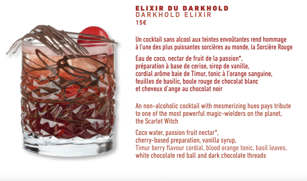 Un verre de cocktail rouge avec de la glace, garni d'une cerise et de décorations en chocolat, servi au Skyline Bar de l'hôtel New York The Art Of Marvel, à côté d'une description de la boisson sans alcool "Elixir du Darkhold".