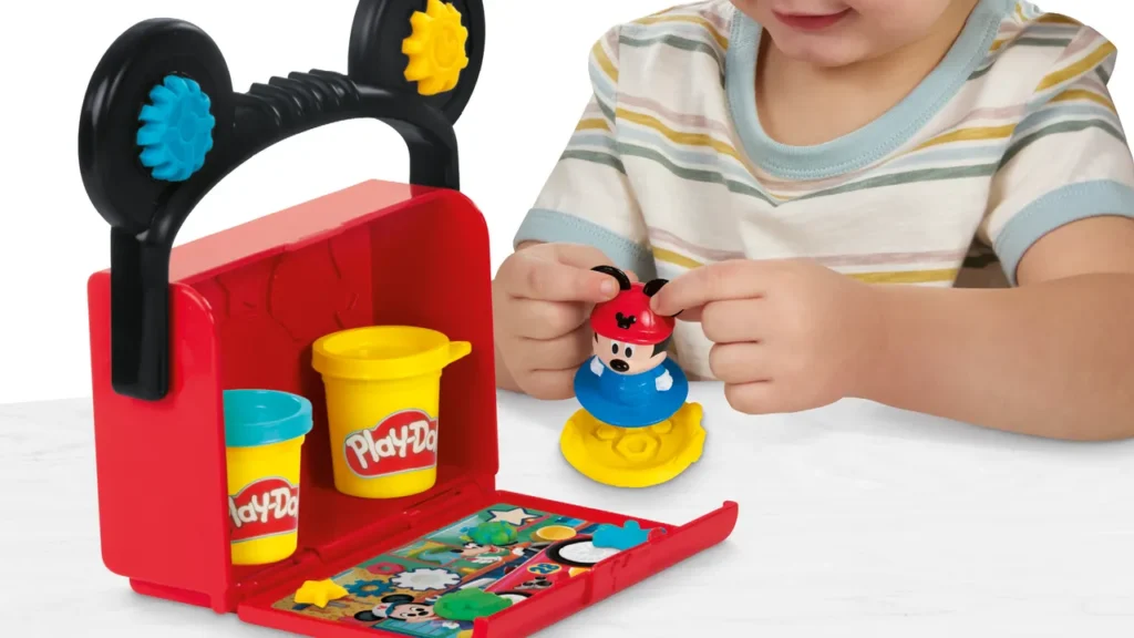 Un enfant joue avec un ensemble Hasbro Disney Play-Doh sur le thème de Mickey Mouse Clubhouse, comprenant des récipients, des moules et des accessoires Play-Doh sur une surface blanche.