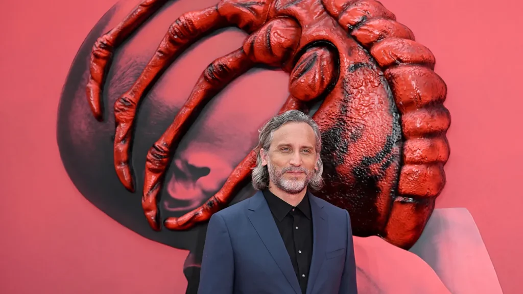 Un homme vêtu d'un costume bleu se tient devant une toile de fond rouge sur laquelle est fixée la tête d'une grande créature extraterrestre de la franchise Alien.