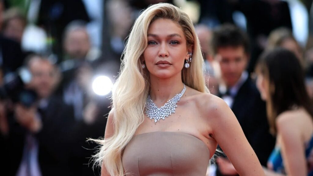 Gigi Hadid, aux longs cheveux blonds, est éblouissante dans une robe beige sans bretelles et un collier de diamants lors d'un événement formel, incarnant Raiponce dans le remake du film en live-action de Disney, tandis que les photographes capturent le moment.
