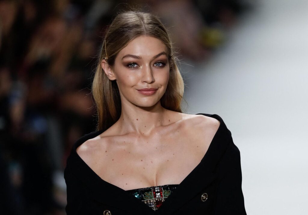 Une femme aux longs cheveux châtains clairs marche sur un podium dans une tenue noire décolletée, regardant la caméra avec assurance. Son style rappelle celui de Gigi Hadid ou de Raiponce dans un remake d'un film de Disney en prises de vues réelles.