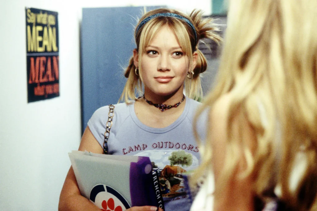 Une adolescente aux cheveux blonds et aux nattes se tient dans un couloir en tenant un dossier, face à une autre fille. Une affiche "MEAN" est visible sur le mur derrière elle, évoquant les vibrations classiques de Lizzie McGuire, parfaites pour un reboot d'Hilary Duff.