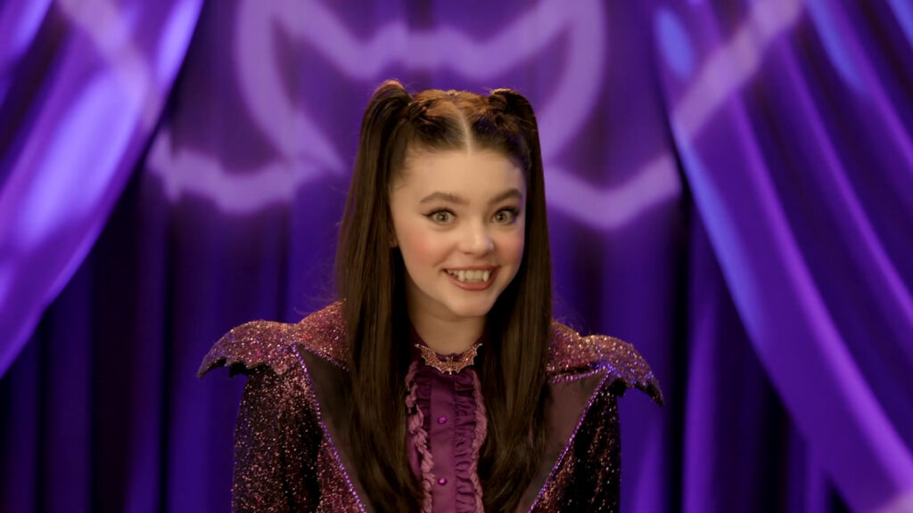 Une jeune femme aux longs cheveux noirs en nattes, vêtue d'un costume violet étincelant inspiré de Vampirina, sourit devant un fond violet avec un motif en forme de chauve-souris.