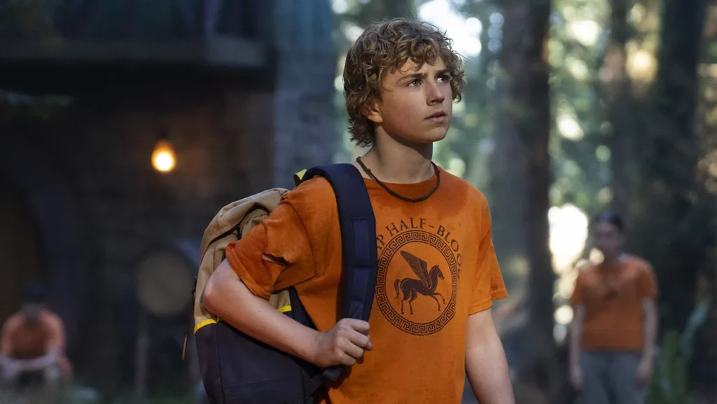Un garçon aux cheveux bouclés portant un T-shirt orange "Camp Half-Blood" et un sac à dos se tient à l'extérieur dans une zone boisée, regardant vers l'avant - laissant présager de nouvelles aventures pour Percy Jackson Saison 2 sur Disney+ après la dernière bande-annonce.