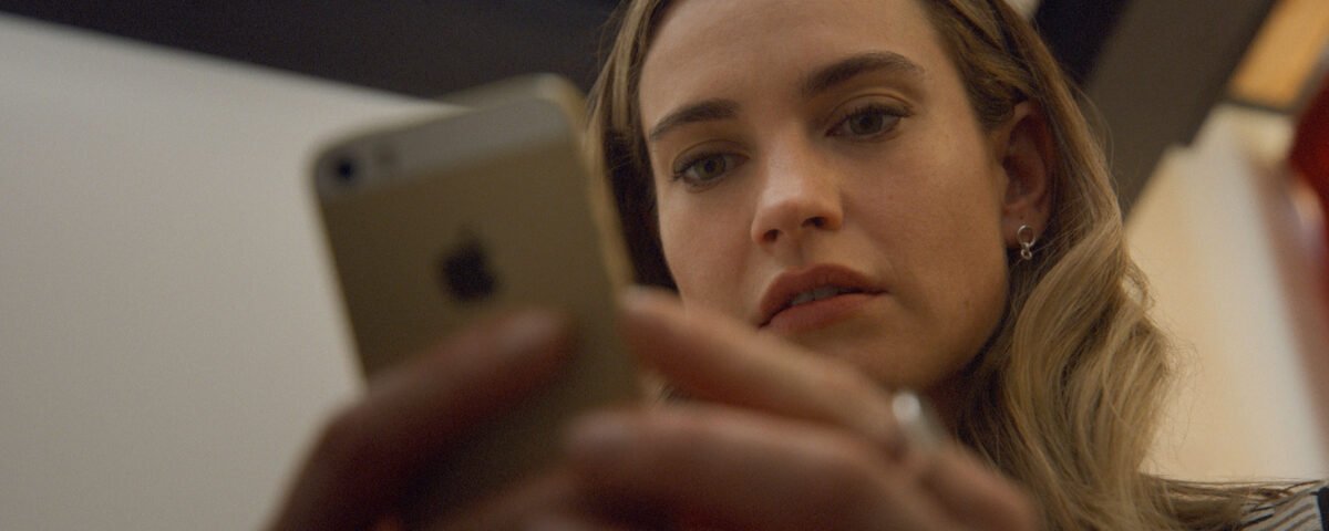 Lily James, les cheveux longs, regarde attentivement un smartphone qu'elle tient à deux mains, peut-être prête à swiper Disney+ ou à lire le biopic créatrice de Bumble.