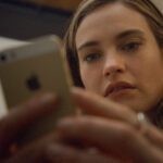 Lily James, les cheveux longs, regarde attentivement un smartphone qu'elle tient à deux mains, peut-être prête à swiper Disney+ ou à lire le biopic créatrice de Bumble.