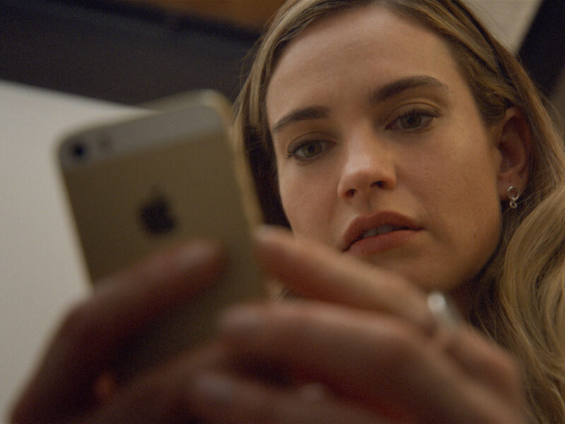 Lily James, les cheveux longs, regarde attentivement un smartphone qu'elle tient à deux mains, peut-être prête à swiper Disney+ ou à lire le biopic créatrice de Bumble.