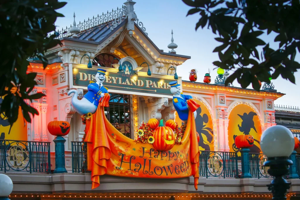 Entrée de Disneyland Paris décorée pour Halloween 2025 avec des lumières orange, des citrouilles, des guirlandes d'automne, des silhouettes et deux fantômes tenant une banderole "Happy Halloween". La parade de Mickey ajoute une touche festive à cette scène sinistre.