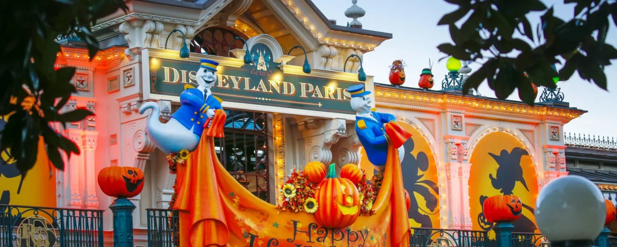 Entrée de Disneyland Paris décorée pour Halloween 2025 avec des lumières orange, des citrouilles, des guirlandes d'automne, des silhouettes et deux fantômes tenant une banderole "Happy Halloween". La parade de Mickey ajoute une touche festive à cette scène sinistre.