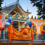 Entrée de Disneyland Paris décorée pour Halloween 2025 avec des lumières orange, des citrouilles, des guirlandes d'automne, des silhouettes et deux fantômes tenant une banderole "Happy Halloween". La parade de Mickey ajoute une touche festive à cette scène sinistre.