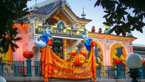 Entrée de Disneyland Paris décorée pour Halloween 2025 avec des lumières orange, des citrouilles, des guirlandes d'automne, des silhouettes et deux fantômes tenant une banderole "Happy Halloween". La parade de Mickey ajoute une touche festive à cette scène sinistre.