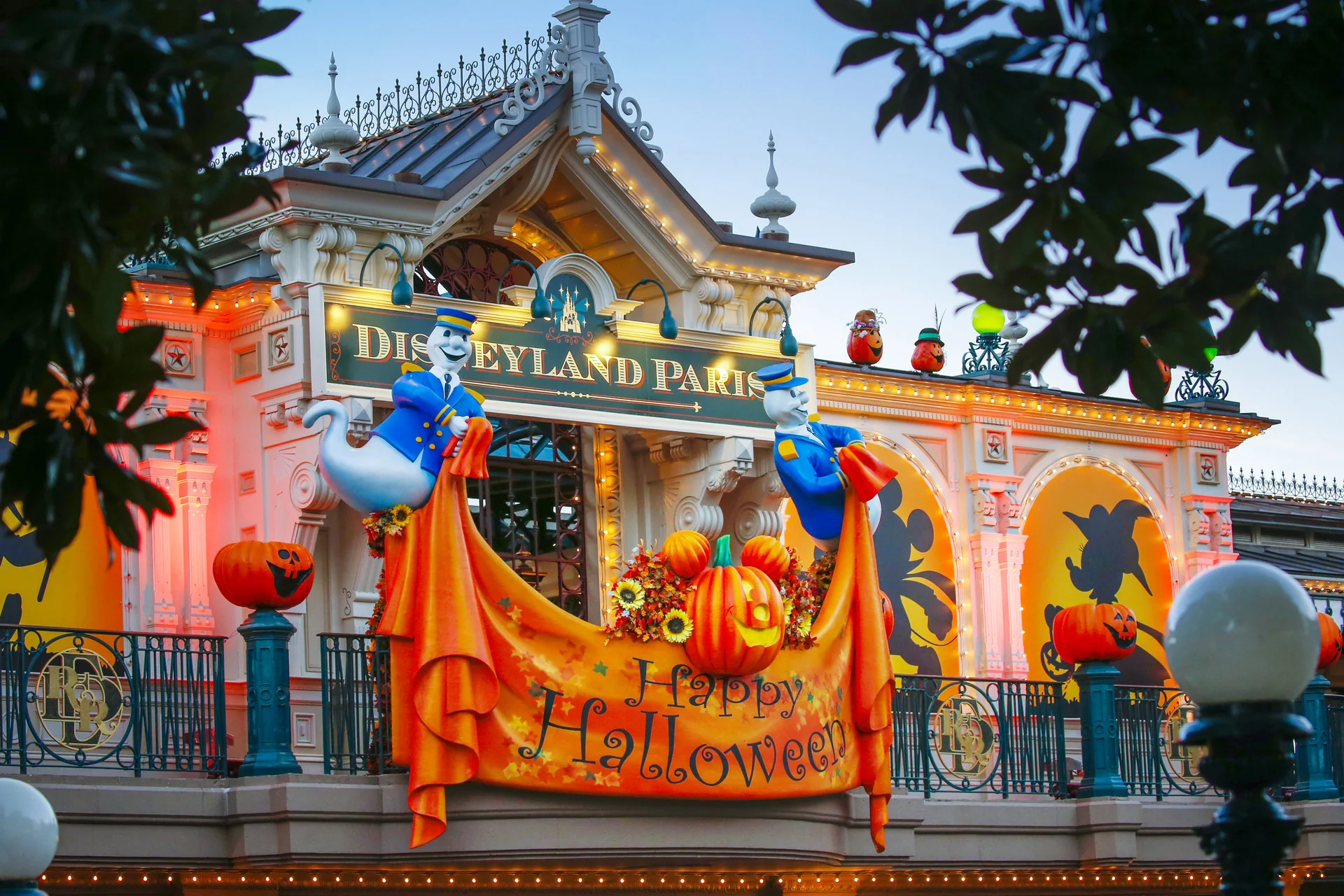 Entrée de Disneyland Paris décorée pour Halloween 2025 avec des lumières orange, des citrouilles, des guirlandes d'automne, des silhouettes et deux fantômes tenant une banderole "Happy Halloween". La parade de Mickey ajoute une touche festive à cette scène sinistre.