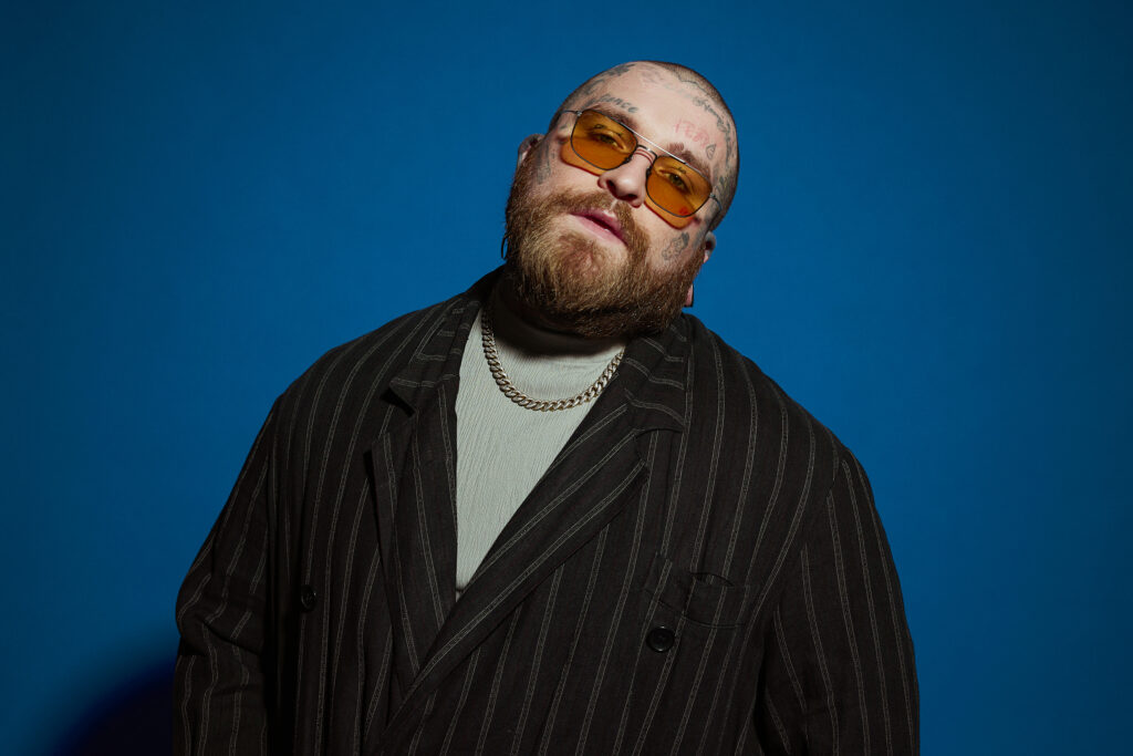 Un barbu au visage tatoué, portant des lunettes de soleil jaunes, un manteau noir rayé, un haut gris et un collier à chaîne, pose sur un fond bleu dans un style inspiré de Pharrell.