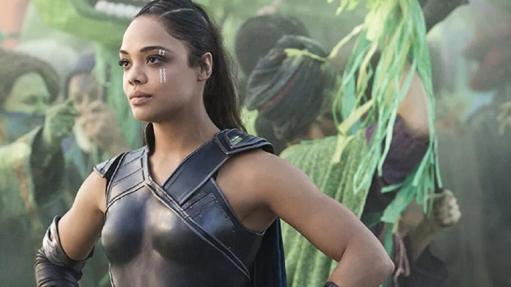 Une femme en armure noire et au visage peint se tient avec assurance, les mains sur les hanches, incarnant un personnage puissant rappelant Valkyrie Marvel, entourée de personnes vêtues de costumes verts.