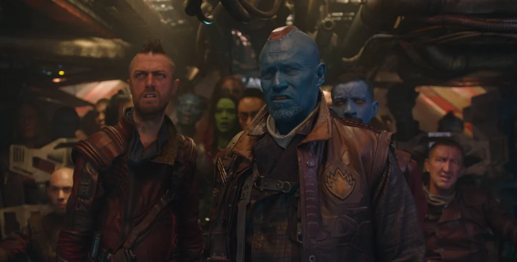 Un groupe de personnes, dont un homme à la peau bleue vêtu d'une veste rouge, se tient debout à l'intérieur d'un vaisseau spatial faiblement éclairé, d'apparence industrielle, qui fait clairement partie des célèbres Ravageurs Marvel.