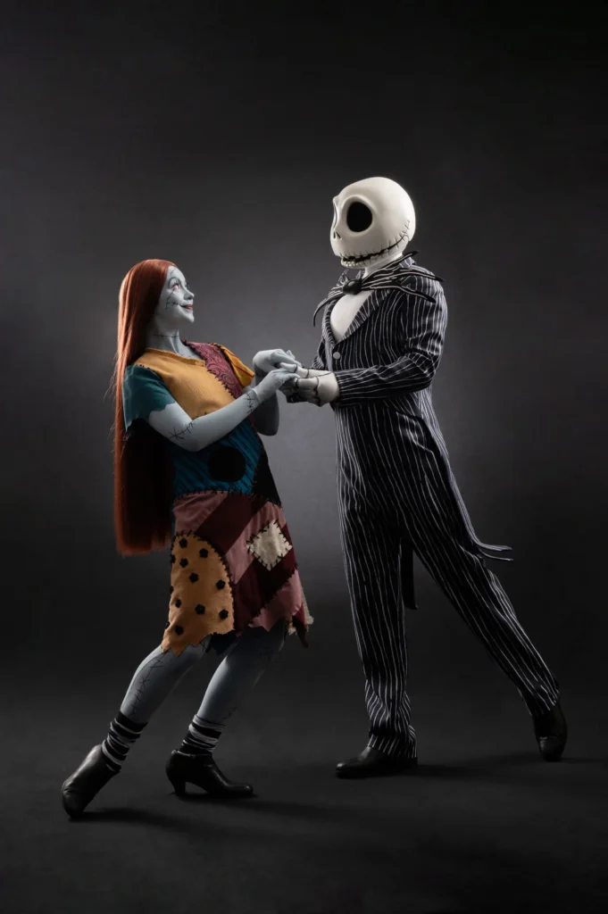 Deux personnes en costumes de Sally et Jack Skellington de "The Nightmare Before Christmas" se tiennent la main sur un fond sombre, prêtes pour la célébration d'Halloween Disney 2025 à Disneyland Paris.