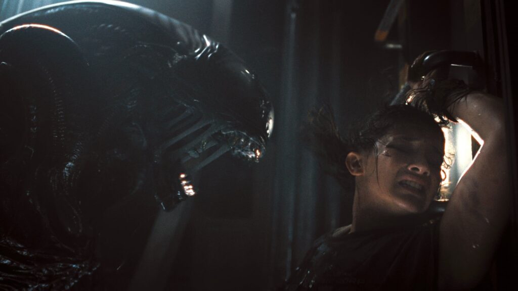 Une femme se presse contre un mur, effrayée, tandis qu'une grande créature extraterrestre menaçante - rappelant Alien Romulus - s'agite près d'elle dans un espace faiblement éclairé. Ce film illustre bien le style de suspense que le réalisateur Fede Álvarez apporte à la franchise Disney.