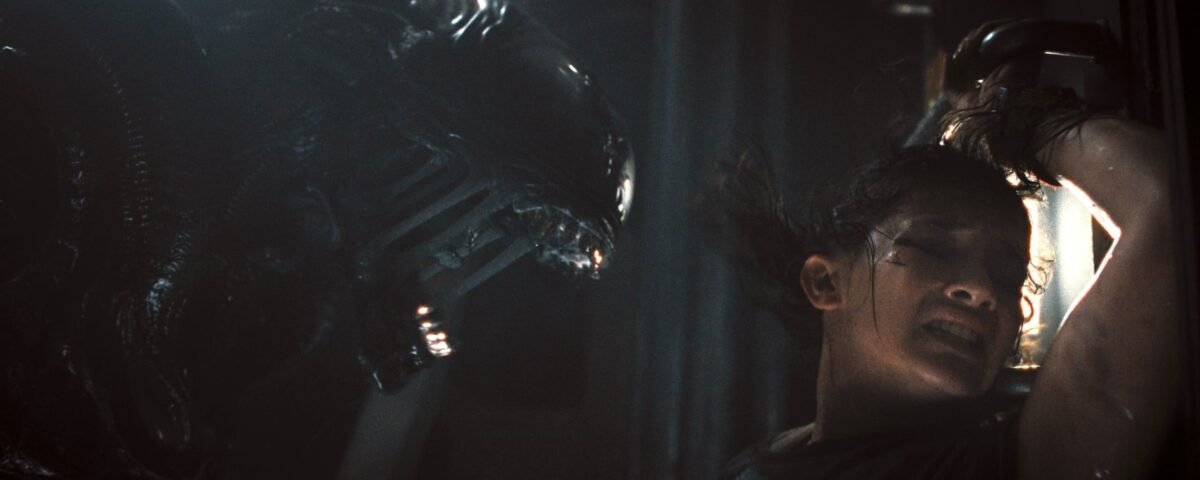 Une femme se presse contre un mur, effrayée, tandis qu'une grande créature extraterrestre menaçante - rappelant Alien Romulus - s'agite près d'elle dans un espace faiblement éclairé. Ce film illustre bien le style de suspense que le réalisateur Fede Álvarez apporte à la franchise Disney.