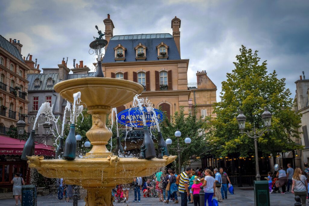 Une fontaine décorative avec des bouteilles de champagne pulvérise de l'eau sur une place très fréquentée de Disneyland Paris, entourée de bâtiments et d'arbres de style parisien, où les gens choisissent leur billet et se rassemblent pour profiter de l'atmosphère animée.