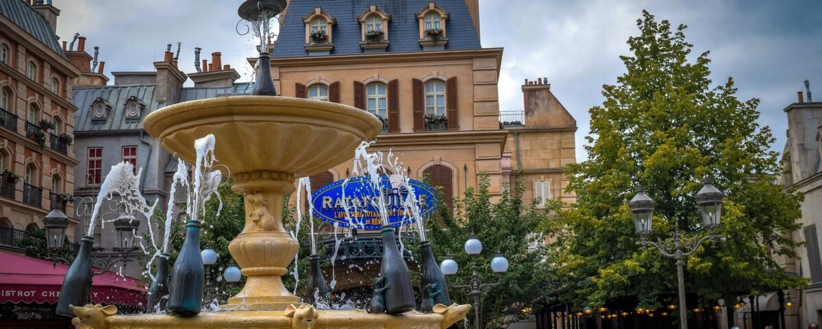 Une fontaine décorative avec des bouteilles de champagne pulvérise de l'eau sur une place très fréquentée de Disneyland Paris, entourée de bâtiments et d'arbres de style parisien, où les gens choisissent leur billet et se rassemblent pour profiter de l'atmosphère animée.