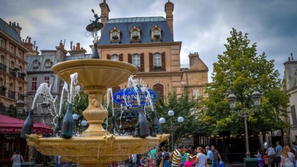 Une fontaine décorative avec des bouteilles de champagne pulvérise de l'eau sur une place très fréquentée de Disneyland Paris, entourée de bâtiments et d'arbres de style parisien, où les gens choisissent leur billet et se rassemblent pour profiter de l'atmosphère animée.