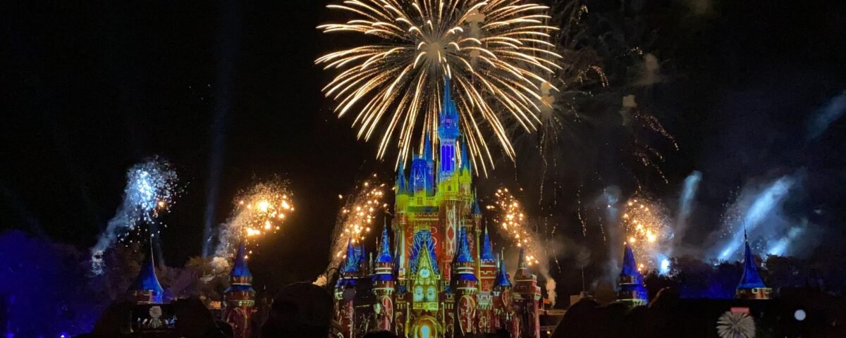 Des feux d'artifice illuminent le ciel nocturne au-dessus d'un château illuminé dans l'un des célèbres parcs d'attractions. Des silhouettes de spectateurs capturent l'instant sur leur téléphone, grâce aux récentes avancées technologiques.