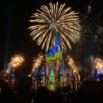 Des feux d'artifice illuminent le ciel nocturne au-dessus d'un château illuminé dans l'un des célèbres parcs d'attractions. Des silhouettes de spectateurs capturent l'instant sur leur téléphone, grâce aux récentes avancées technologiques.