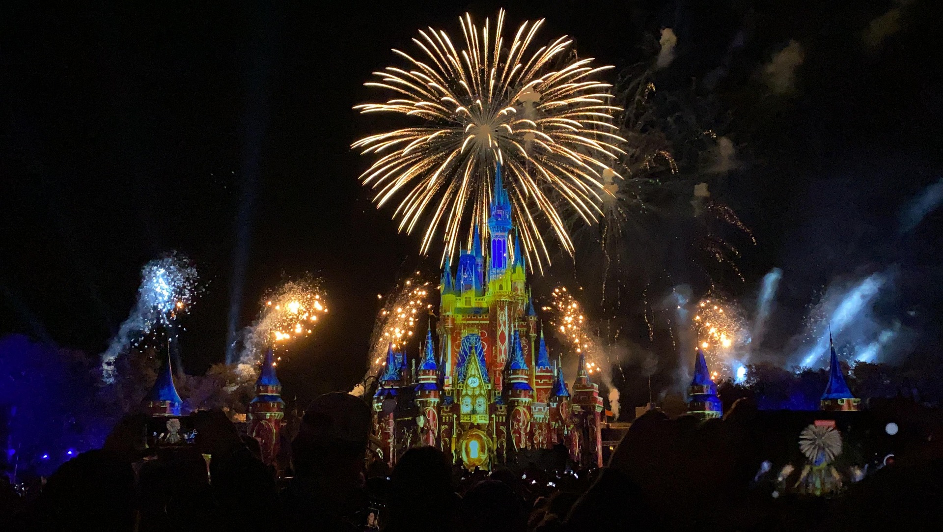 Des feux d'artifice illuminent le ciel nocturne au-dessus d'un château illuminé dans l'un des célèbres parcs d'attractions. Des silhouettes de spectateurs capturent l'instant sur leur téléphone, grâce aux récentes avancées technologiques.