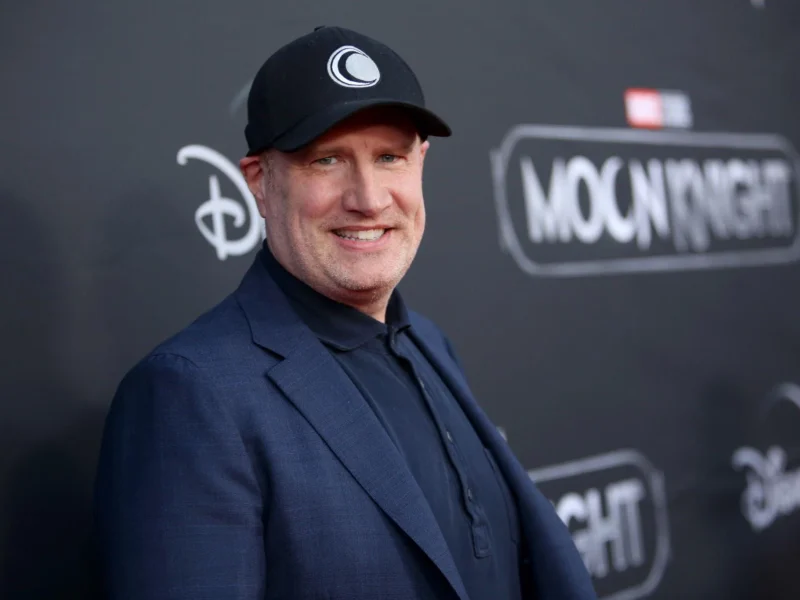 Un homme vêtu d'un blazer sombre et d'une casquette noire se tient devant une toile de fond de l'événement "Moon Knight" sur laquelle figurent les logos de Disney+ et de Marvel Studios.