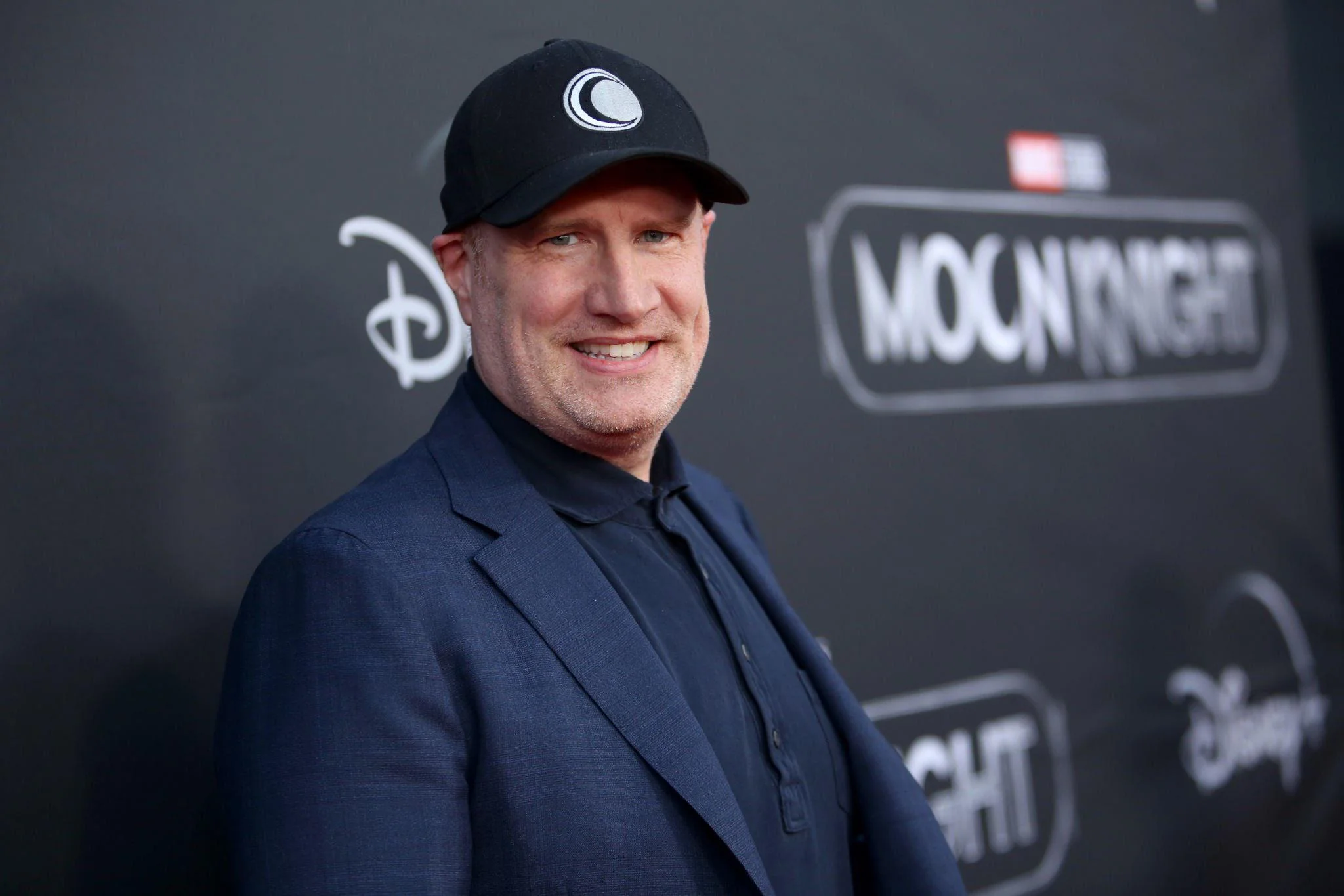 Un homme vêtu d'un blazer sombre et d'une casquette noire se tient devant une toile de fond de l'événement "Moon Knight" sur laquelle figurent les logos de Disney+ et de Marvel Studios.