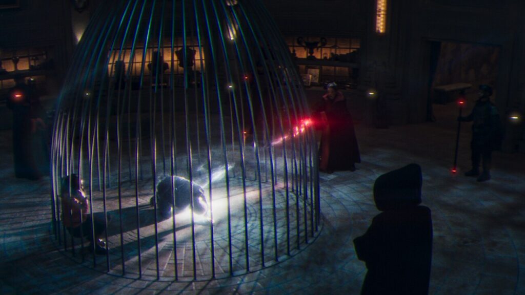 Une personne est agenouillée à l'intérieur d'une grande cage métallique, éclairée par des faisceaux de lumière provenant de personnages à l'extérieur, dans une pièce faiblement éclairée où se trouvent plusieurs spectateurs - une scène qui rappelle l'atmosphère sinistre de Marvel dans Werewolf By Night.