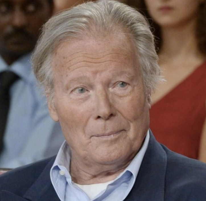 Un homme âgé aux cheveux gris, portant un blazer sombre et une chemise bleu clair, est assis à l'intérieur, tandis que d'autres personnes à l'arrière-plan flou semblent converser. L'atmosphère suggère un cadre dans lequel chacun est encouragé à être prêt pour la suite.