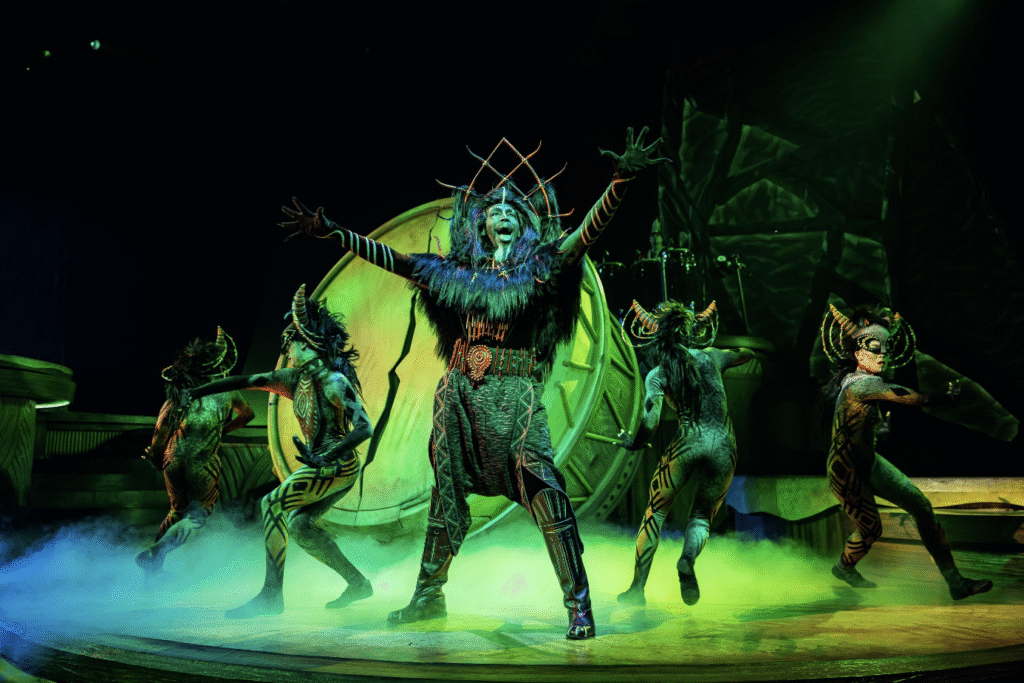 Des artistes vêtus de costumes élaborés, inspirés des animaux, dansent sur une scène brumeuse et éclairée de couleurs vives lors d'une production théâtrale.
