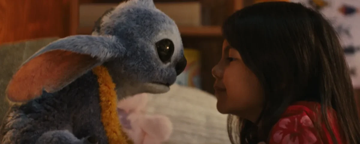 Une jeune fille fait face à une créature extraterrestre bleue en peluche avec de grandes oreilles et des yeux sombres dans un décor intérieur confortable, rappelant le film bien-aimé Lilo & Stitch.