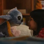 Une jeune fille fait face à une créature extraterrestre bleue en peluche avec de grandes oreilles et des yeux sombres dans un décor intérieur confortable, rappelant le film bien-aimé Lilo & Stitch.