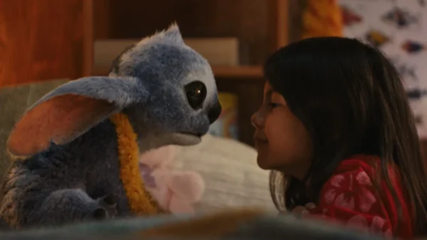 Une jeune fille fait face à une créature extraterrestre bleue en peluche avec de grandes oreilles et des yeux sombres dans un décor intérieur confortable, rappelant le film bien-aimé Lilo & Stitch.