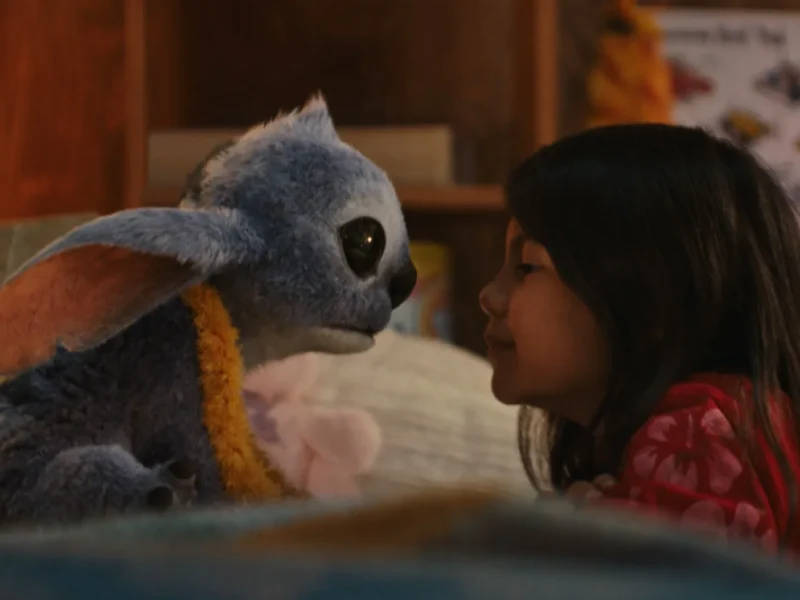 Une jeune fille fait face à une créature extraterrestre bleue en peluche avec de grandes oreilles et des yeux sombres dans un décor intérieur confortable, rappelant le film bien-aimé Lilo & Stitch.