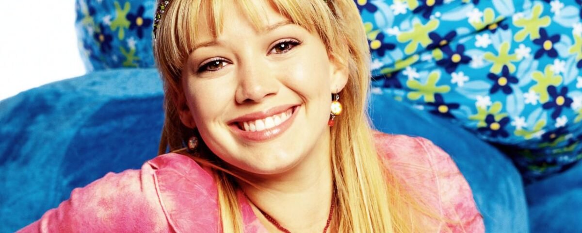 Une jeune femme aux longs cheveux blonds, vêtue d'un haut rose et d'un bandeau orné de bijoux, sourit à la caméra. Des coussins bleus à motifs floraux se trouvent à l'arrière-plan, rappelant le style joyeux de Lizzie McGuire dans le reboot d'Hilary Duff sur Disney.