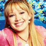 Une jeune femme aux longs cheveux blonds, vêtue d'un haut rose et d'un bandeau orné de bijoux, sourit à la caméra. Des coussins bleus à motifs floraux se trouvent à l'arrière-plan, rappelant le style joyeux de Lizzie McGuire dans le reboot d'Hilary Duff sur Disney.