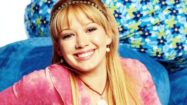 Une jeune femme aux longs cheveux blonds, vêtue d'un haut rose et d'un bandeau orné de bijoux, sourit à la caméra. Des coussins bleus à motifs floraux se trouvent à l'arrière-plan, rappelant le style joyeux de Lizzie McGuire dans le reboot d'Hilary Duff sur Disney.