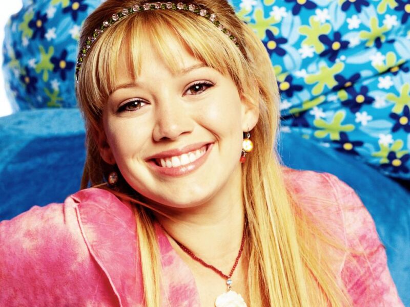 Une jeune femme aux longs cheveux blonds, vêtue d'un haut rose et d'un bandeau orné de bijoux, sourit à la caméra. Des coussins bleus à motifs floraux se trouvent à l'arrière-plan, rappelant le style joyeux de Lizzie McGuire dans le reboot d'Hilary Duff sur Disney.