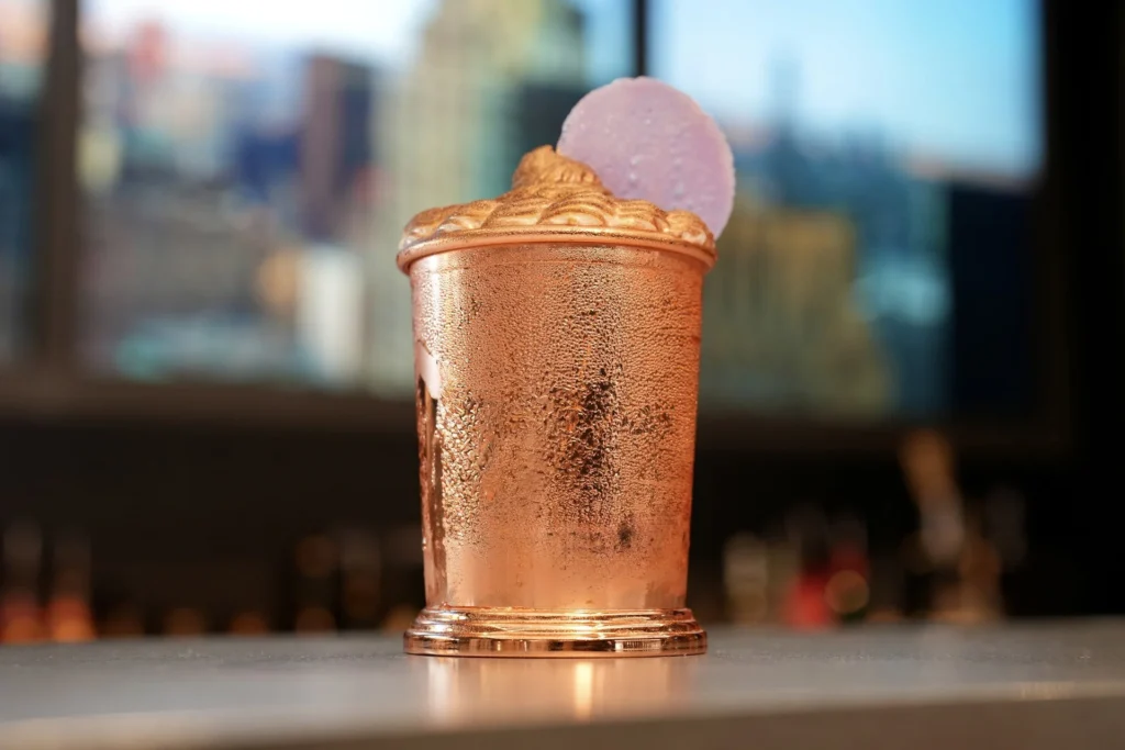 Une tasse en cuivre remplie d'une boisson mousseuse et surmontée d'une garniture ronde violette trône sur le comptoir du Skyline Bar, à l'hôtel New York, sur fond de paysage urbain flou - parfait pour les amateurs de cocktails ou de Super Héros.