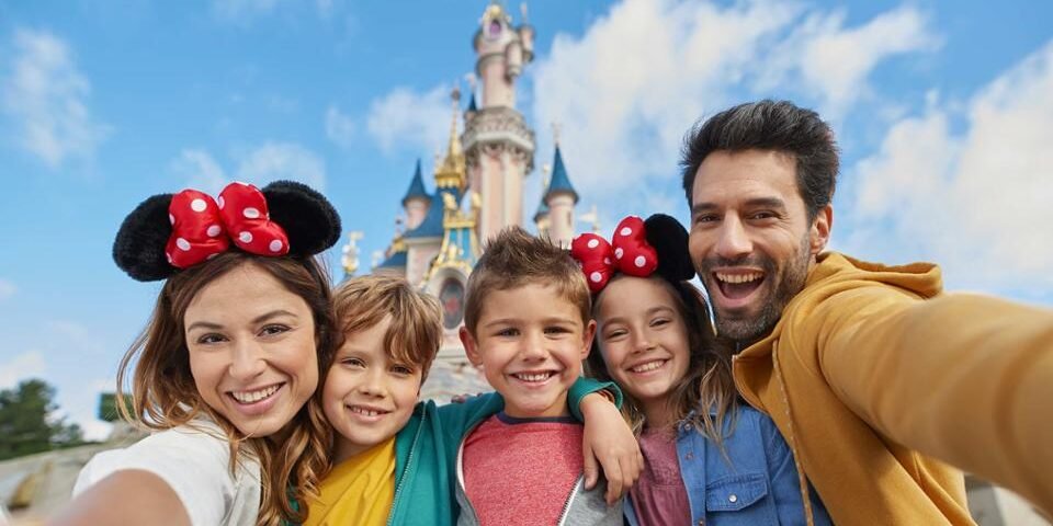 Une famille de cinq personnes prend un selfie devant un château à Disneyland Paris, avec deux enfants portant des bandeaux d'oreilles Minnie Mouse - excités après avoir trouvé des façons amusantes d'occuper l'enfant file d'attente.