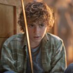 Un jeune garçon aux cheveux bouclés, vêtu d'une chemise à carreaux verte, est assis par terre à l'intérieur, tenant une épée droite devant son visage - ce qui rappelle des scènes de la bande-annonce Percy Jackson Saison 2 sur Disney+.