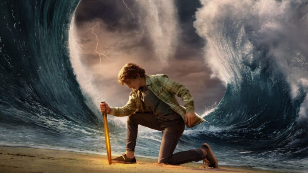 Un jeune homme est agenouillé sur le sable, tenant une épée lumineuse, avec de grandes vagues de l'océan se séparant de façon spectaculaire derrière eux sous un ciel orageux, évoquant le style épique de Percy Jackson et Les Olympiens Saison 2, dont la bande-annonce Disney+ fait actuellement l'apologie.