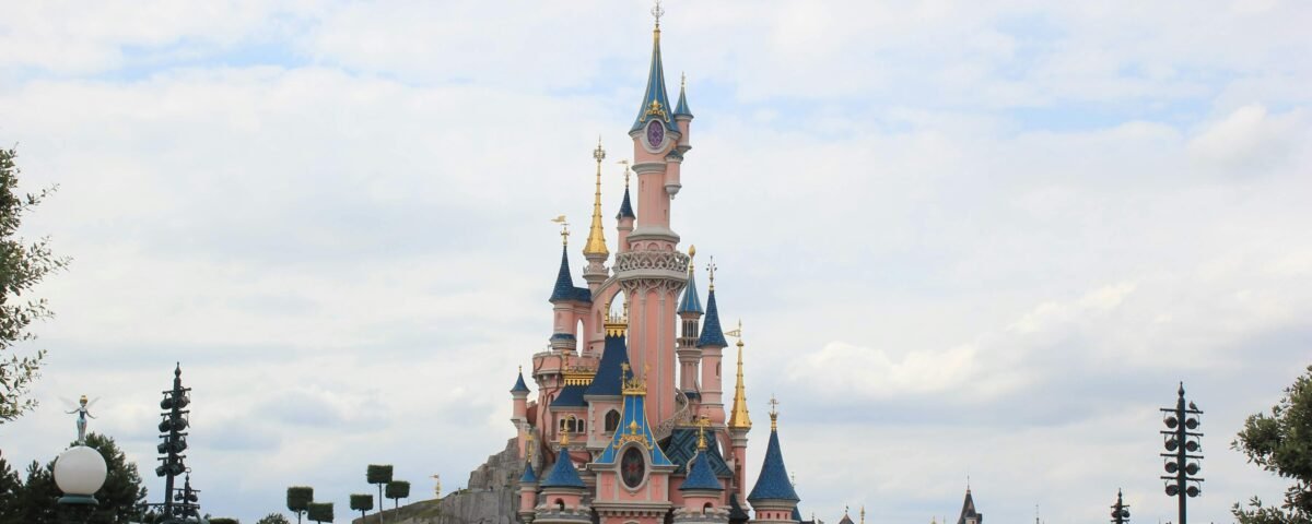Une foule nombreuse se rassemble devant un château de conte de fées rose aux toits bleus, sous un ciel partiellement nuageux à Disneyland Paris, alors que les visiteurs empruntent les files d'attente pour profiter de la magie du parc d'attractions.