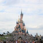 Une foule nombreuse se rassemble devant un château de conte de fées rose aux toits bleus, sous un ciel partiellement nuageux à Disneyland Paris, alors que les visiteurs empruntent les files d'attente pour profiter de la magie du parc d'attractions.