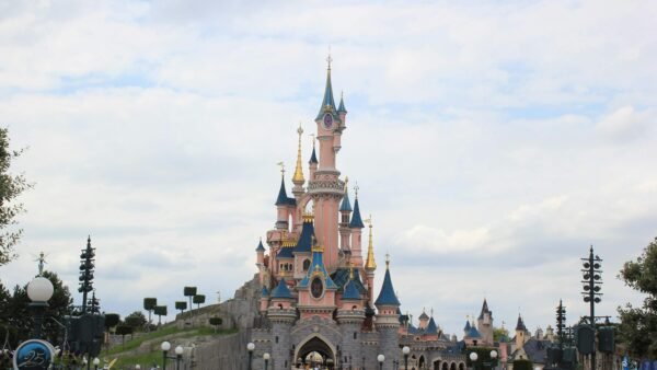 Une foule nombreuse se rassemble devant un château de conte de fées rose aux toits bleus, sous un ciel partiellement nuageux à Disneyland Paris, alors que les visiteurs empruntent les files d'attente pour profiter de la magie du parc d'attractions.